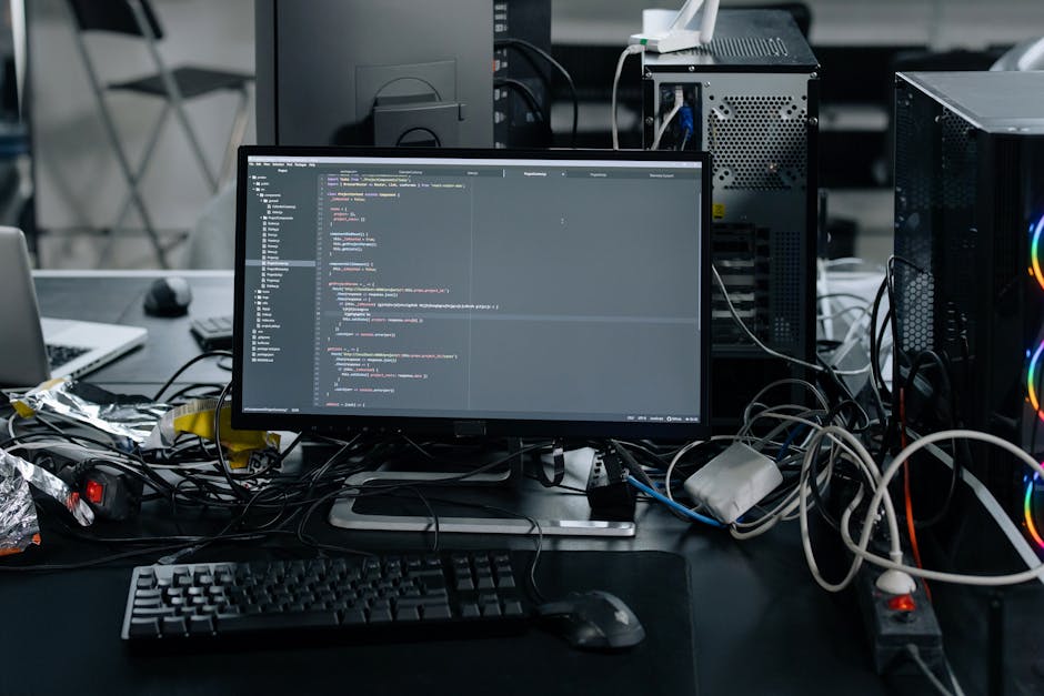 A cluttered workspace featuring a computer monitor displaying lines of code, surrounded by tangled cables, a keyboard, and a mouse on a dark desk. To the right, two desktop towers with visible cooling