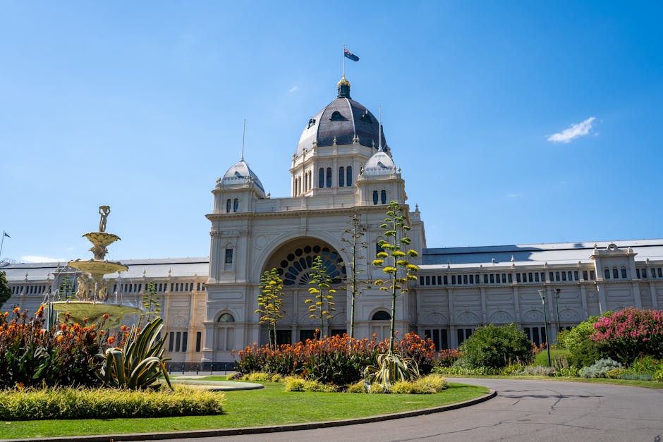 A grand historic building with a large central dome topped with a flag, surrounded by a well-maintained garden. In the foreground, there are vibrant flowering plants and lush green shrubs lining a cur