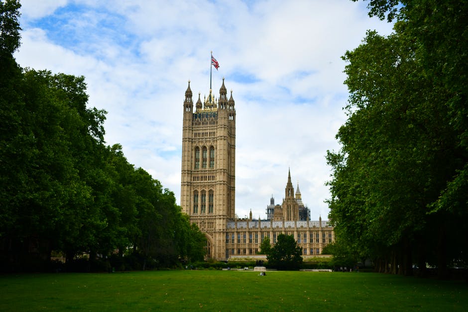 A grand historic building with a large central dome topped with a flag, surrounded by a well-maintained garden. In the foreground, there are vibrant flowering plants and lush green shrubs lining a cur